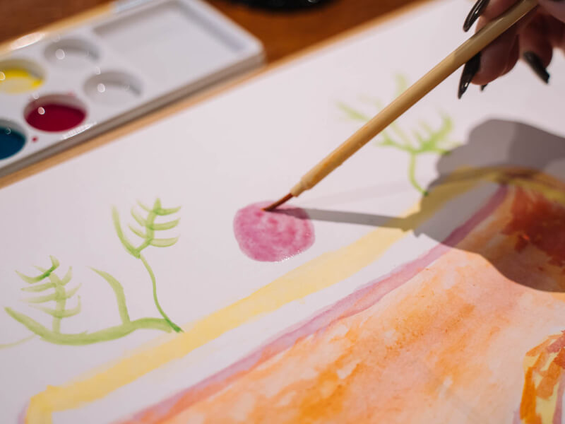 A close up of some paints and a person's hand holding a brush to a white page to begin a water colour